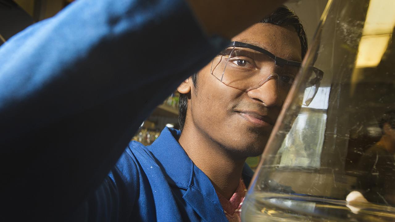 Male student in blue lab coat holding a beaker and looking at the camera