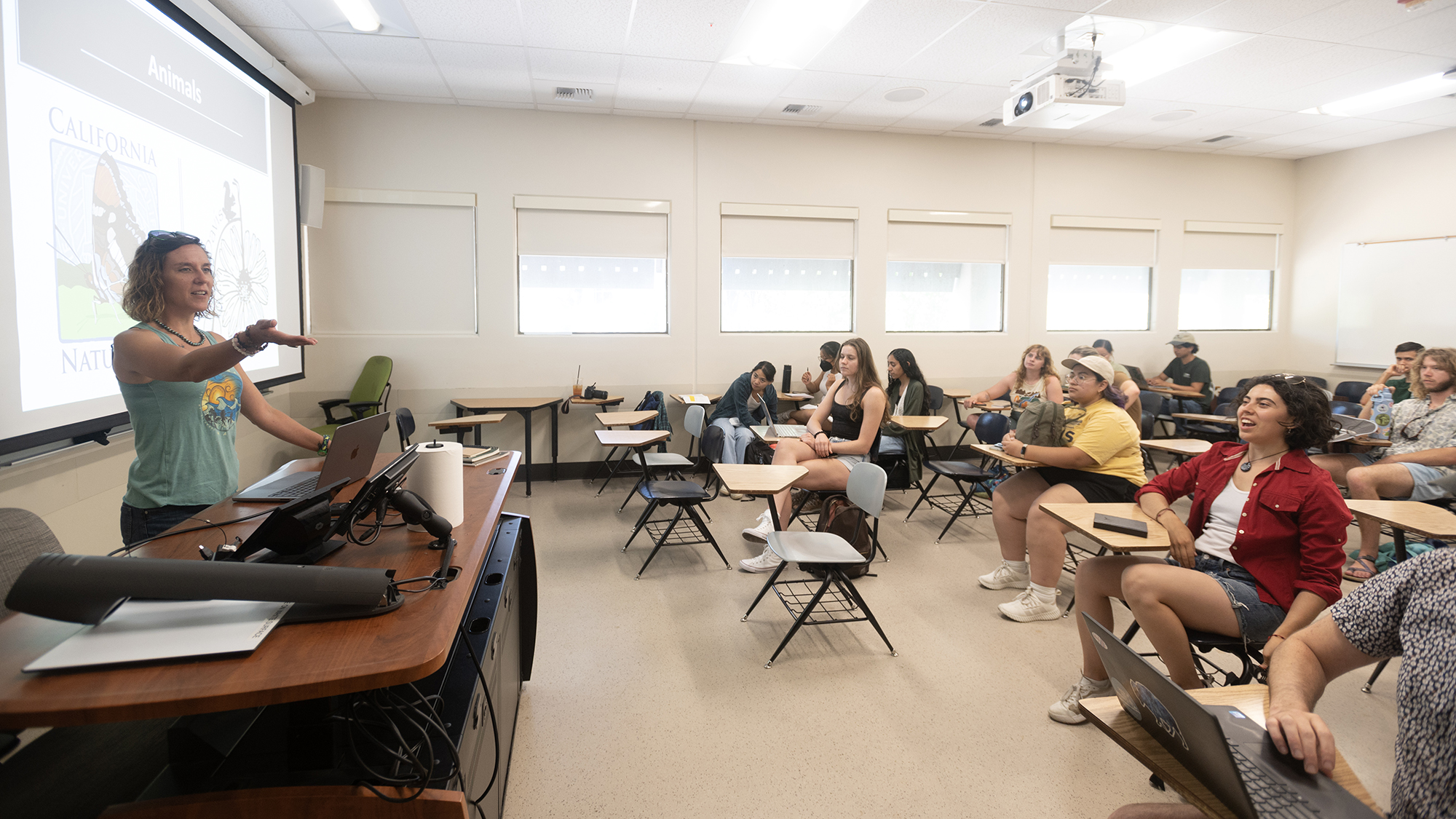 Instructor gestures while speaking at the front of a classroom, engaging a group of students seated at desks; a presentation slide about animals is projected behind her.