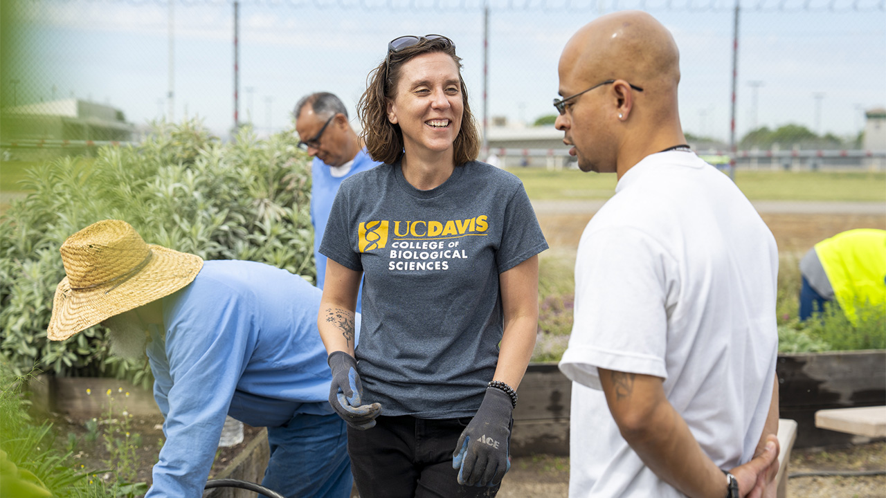 Smiling woman wearing T-shirt with UC Davis logo talks while volunteers garden outdoors