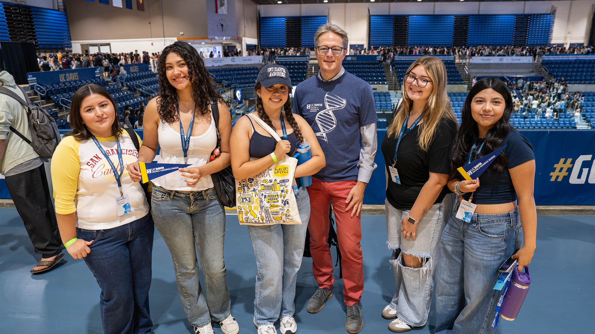 Six smiling young adults standing in a blue indoor stadium concourse, holding bags (photo)
