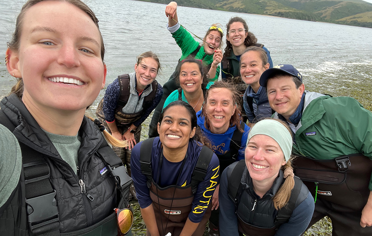Ten smiling people in life jackets pose for a boat selfie on a lake with hills.