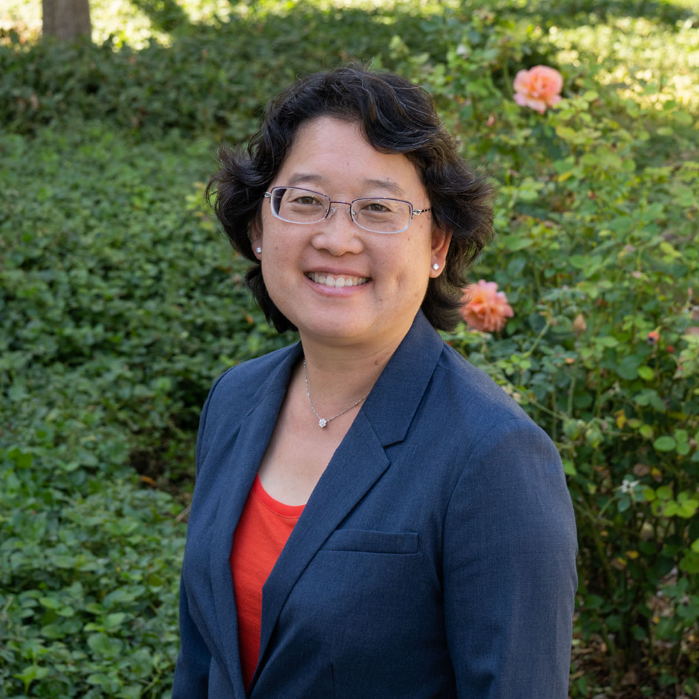 Smiling person wearing glasses, a navy blazer, and an orange top, standing outdoors in front of greenery and blooming roses.