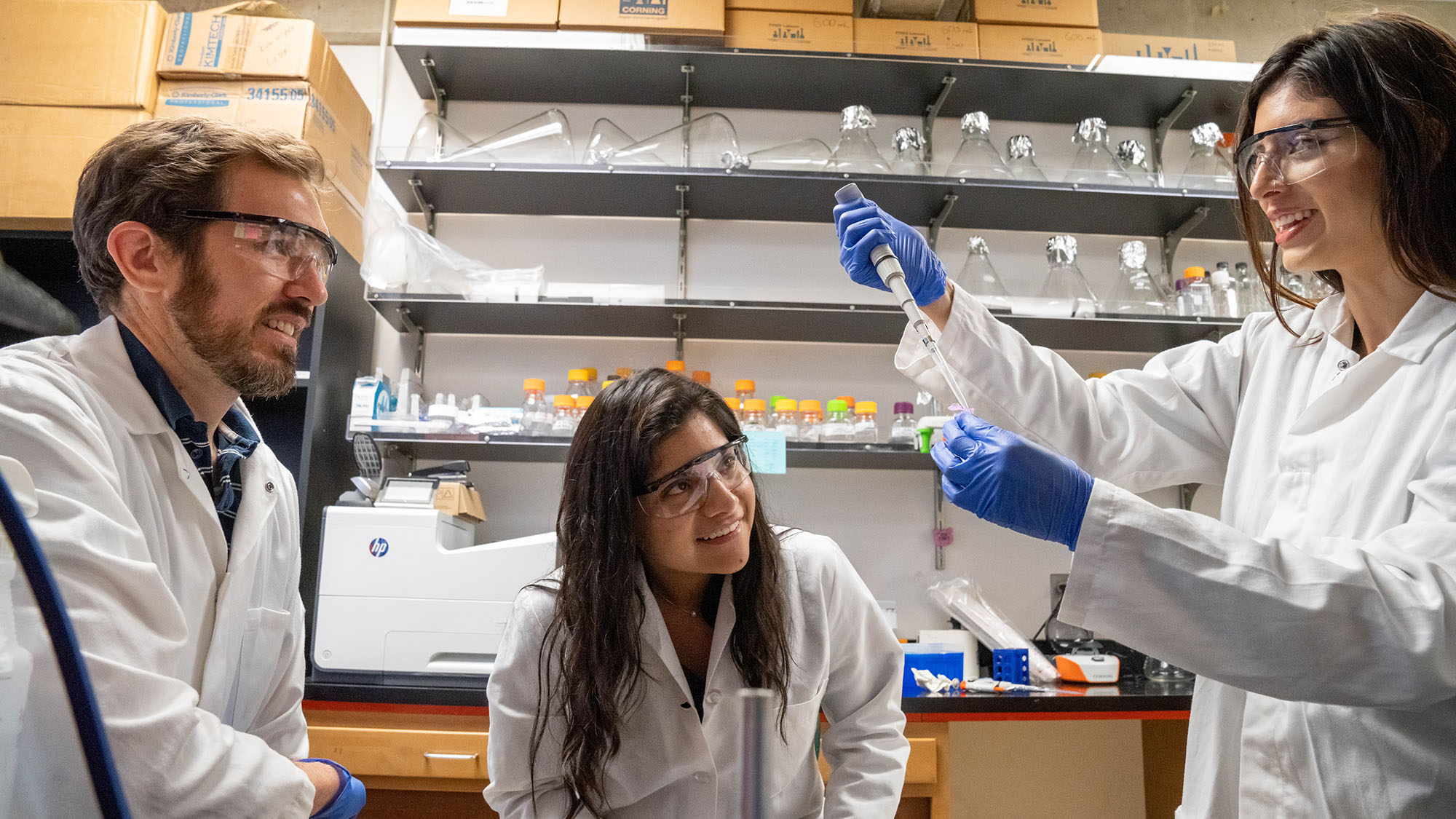 Three people in lab coats and goggles smile as one uses a pipette, with shelves of lab glassware behind them