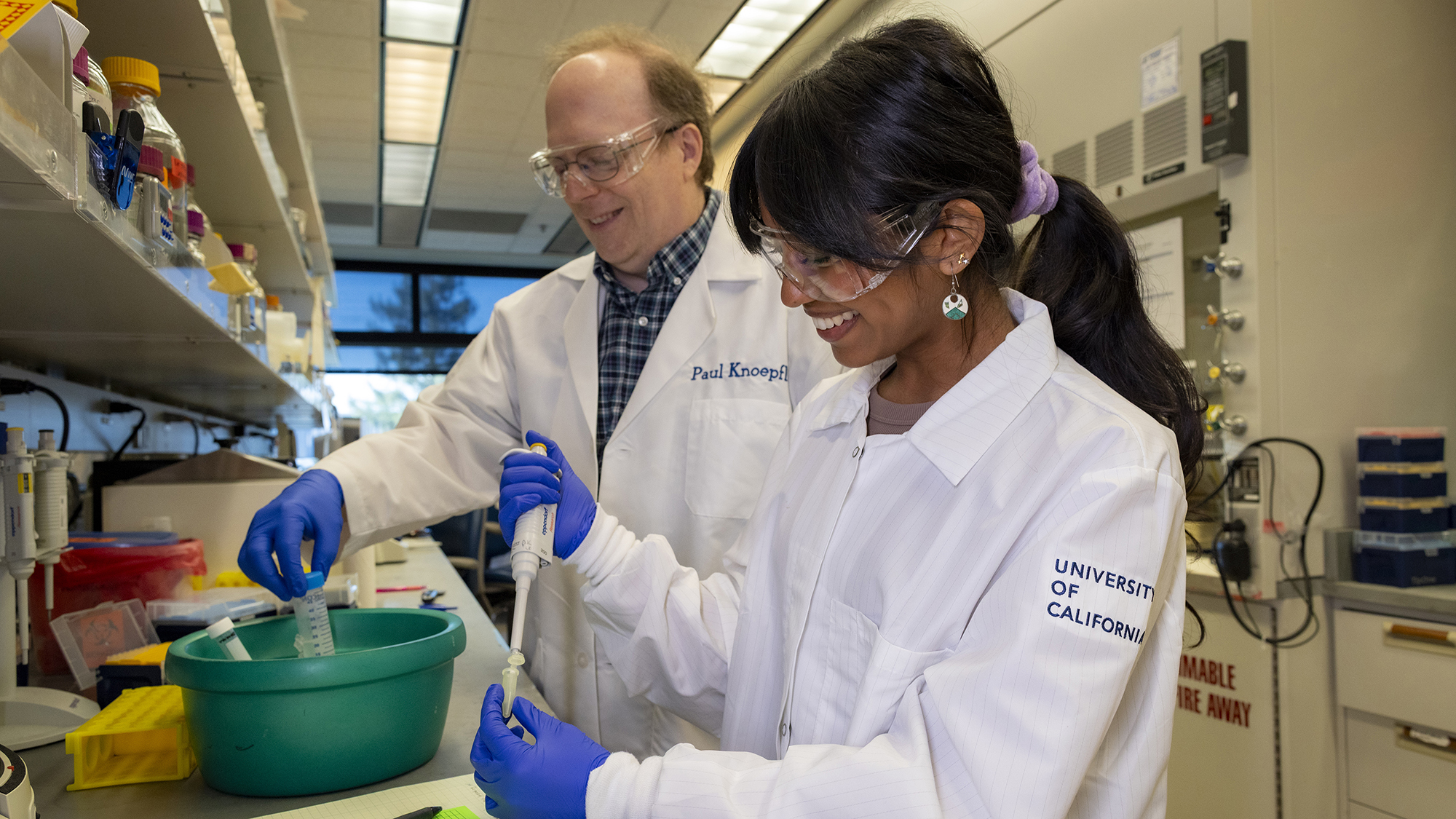 UC Davis student Avantika Gokulnatha and professor Paul Knoepfler work together in a biology lab. Both wear lab coats, safety goggles, and gloves while handling pipettes, surrounded by lab equipment and supplies.