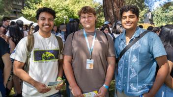 Three smiling students stand together outdoors during orientation, holding campus maps with a crowd of peers behind them.