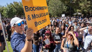 Orientation leader in sunglasses smiles widely while holding a bright “Biochemistry & Molecular Biology” sign before a crowd.