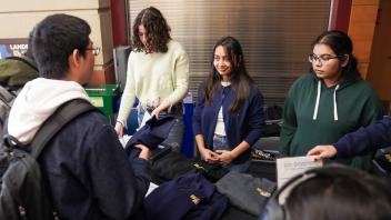 Three young women, wearing casual clothing, stand behind a table displaying UC Davis College of Biological Sciences apparel, assisting a student on the other side of the table.