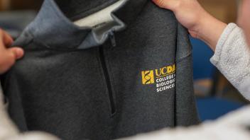 Close-up of a gray UC Davis College of Biological Sciences zip-up sweatshirt held by someone with both hands, focusing on the embroidered logo in white and gold.