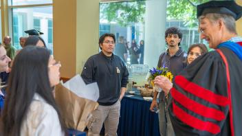 Professor in red-and-black gown hands flowers to smiling students in a lobby
