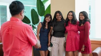 A person photographs four smiling young women posing together near a plant inside a bright UC Davis building. 