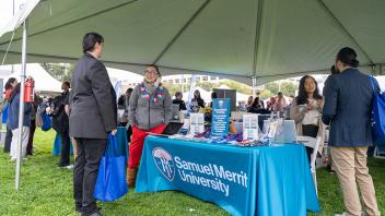 A representative at the Samuel Merritt University booth speaks with attendees under a canopy tent. The table is decorated with a blue tablecloth and brochures, with attendees holding event bags nearby.