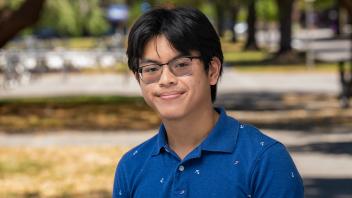 A person in a light blue shirt and glasses standing outside in front of a row of trees