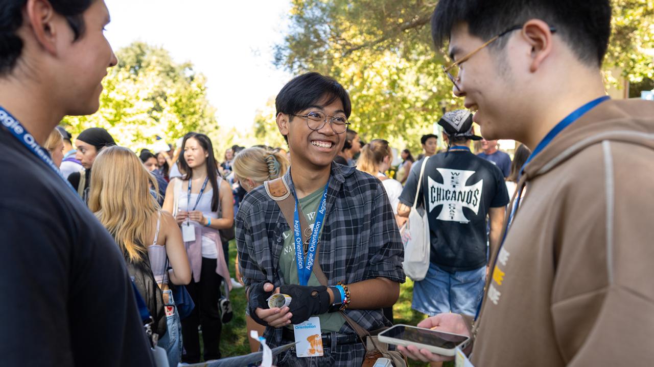 Smiling student wearing glasses chats with two others during an outdoor campus orientation, surrounded by a crowd of peers.