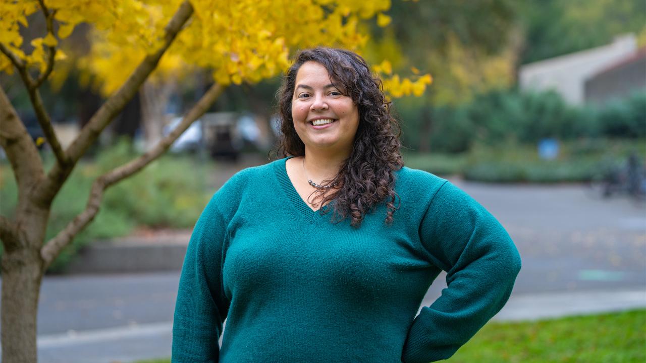 Smiling woman with curly hair, hand on hip, teal sweater outdoors by yellow-leaved tree