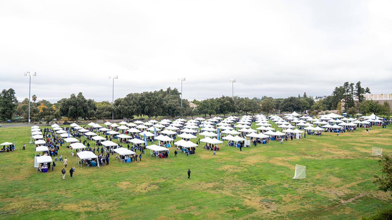 A wide aerial view of a large outdoor event, featuring rows of white canopy tents set up on a grassy field. Attendees can be seen walking between the tents, surrounded by trees and tall light poles in the background.