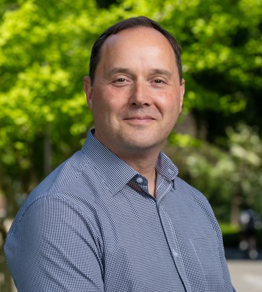A man with short brown hair wearing a blue checkered button-up shirt stands outdoors, smiling slightly at the camera. The background is filled with bright green trees in soft focus.
