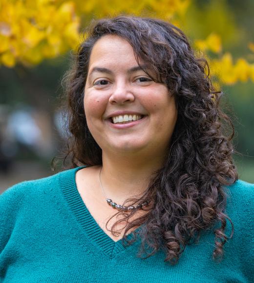 Smiling woman with curly dark hair wearing a teal sweater, photographed outdoors with yellow autumn leaves softly blurred behind her.