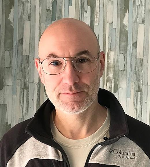 A middle-aged man with a bald head and short gray beard is standing in front of a patterned wall with vertical brushstroke designs in shades of gray, blue, and white. He is wearing large, wire-rimmed glasses and a Columbia Titanium fleece jacket over a beige shirt. He has a neutral expression and is looking directly at the camera.