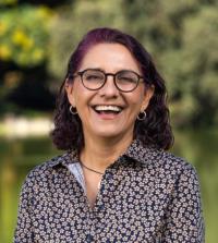 Outdoor headshot of smiling woman with purple hair, glasses, and patterned blouse