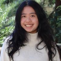A UC Davis student smiles outdoors in front of lush green foliage. She has long black hair and is wearing a cream-colored turtleneck sweater.