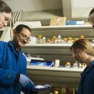 A scientist in a blue lab coat and gloves explains a procedure to three students in a research lab. All are wearing safety goggles and blue UC-branded lab coats, standing in front of shelves lined with bottles and lab supplies.