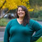 Smiling woman with curly hair, hand on hip, teal sweater outdoors by yellow-leaved tree