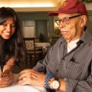 UC Davis student Avantika Gokulnatha shares a phone screen with Jack McGruder, an older man in a red USC hat. They sit at a table smiling together in a warmly lit community space.