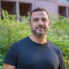 Person standing outdoors, wearing a black t-shirt, short dark hair and beard, greenery and building behind.