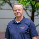 Headshot of a smiling man with short gray hair in a navy polo, blurred trees behind