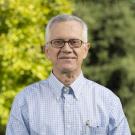 Smiling person with short gray hair and glasses wears a light blue plaid shirt, standing outdoors with trees in the background.