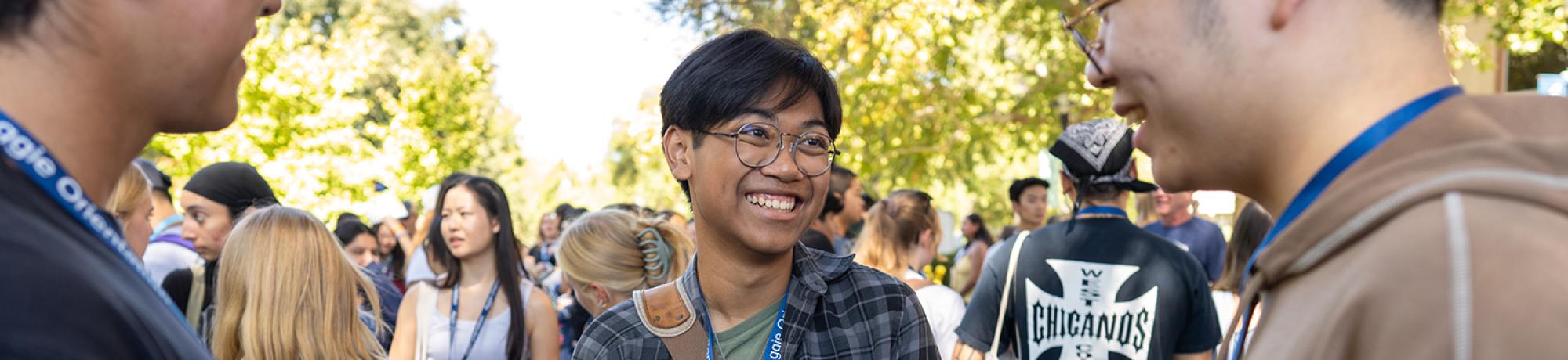 Smiling student wearing glasses chats with two others during an outdoor campus orientation, surrounded by a crowd of peers.