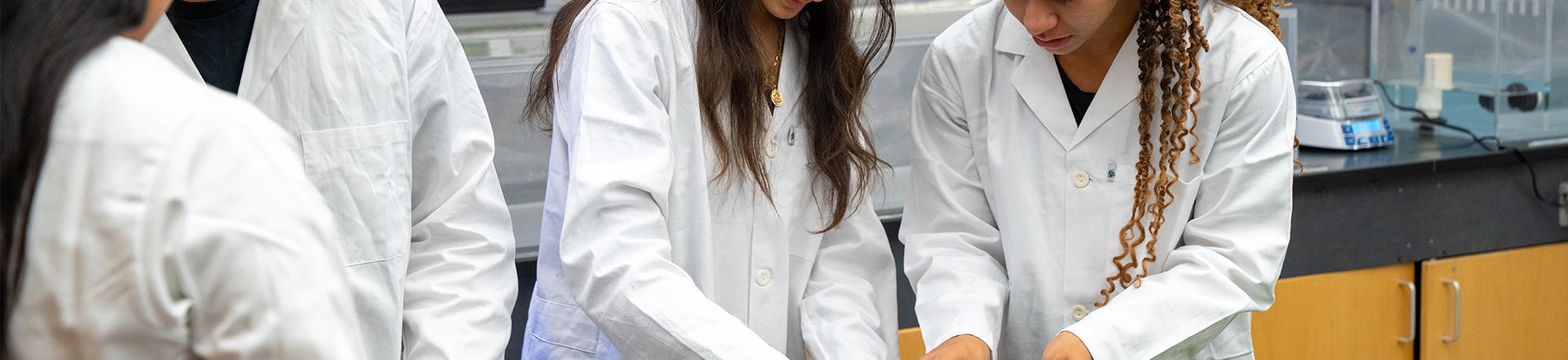 Photo of four students in lab coats examining small green seedlings on a lab bench