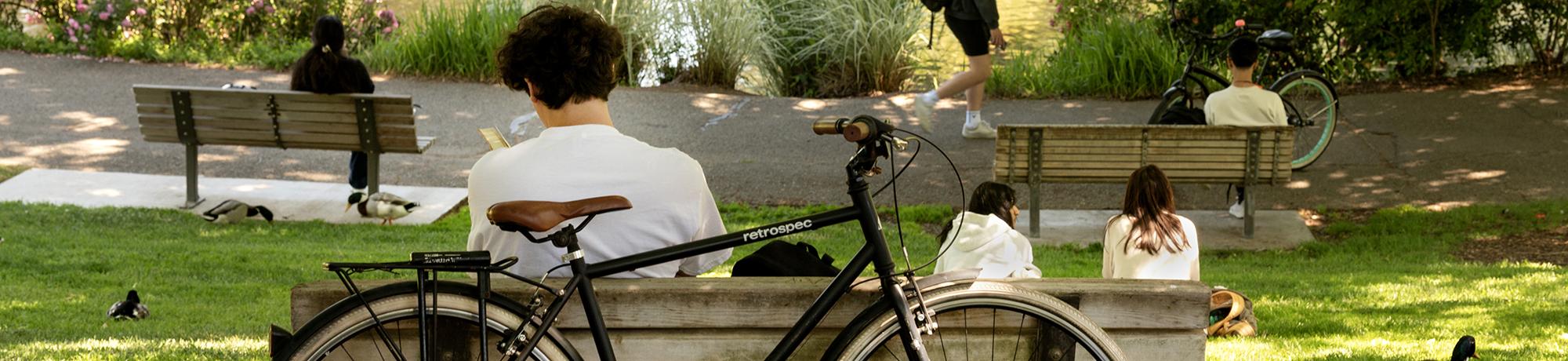  Students relax on benches and grass near a pond at UC Davis, with ducks nearby and bikes parked along the path. One student reads a book with a bicycle in the foreground. Trees and flowers create a peaceful, shaded environment.