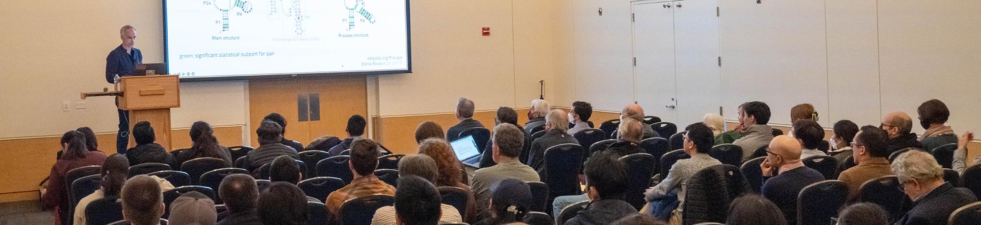Male speaker at the podium in front of projection screen and large audience